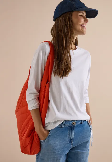 Frau mit weißem Shirt, Jeans, roter Tasche und blauer Kappe, lächelnd seitlich stehend.