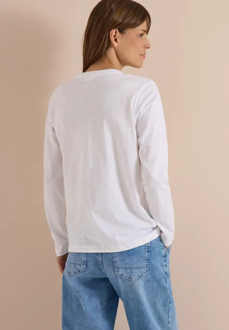 Eine Frau steht im Profil, trägt ein weißes Langarmshirt und blaue Jeans vor hellem Hintergrund.