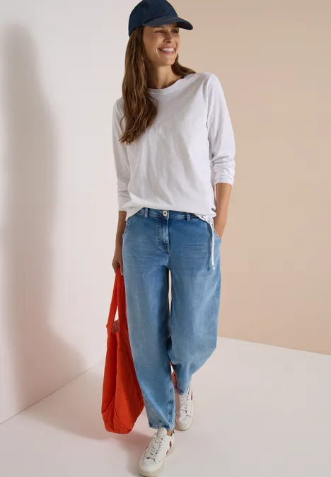 Eine junge Frau in weißen Sneakern, Jeans, weißem Shirt und Schildmütze hält eine rote Tasche.