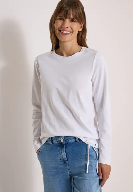 Eine Frau in Jeans und weißem Langarmshirt lächelt vor hellem Hintergrund.