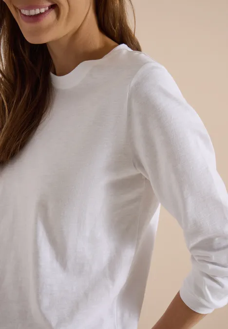 Frau in weißem Langarmshirt, nur Oberkörper und freundliches Lächeln sichtbar.