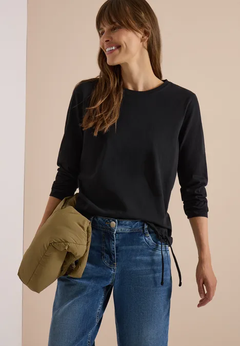 Frau in schwarzem Langarmshirt und Jeans, hält olivgrüne Jacke und lächelt.