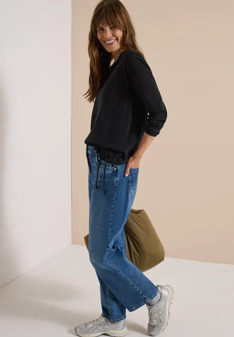 Frau trägt schwarzen Pullover, weite blaue Jeans, weiße Sneaker und hält eine grüne Tasche.