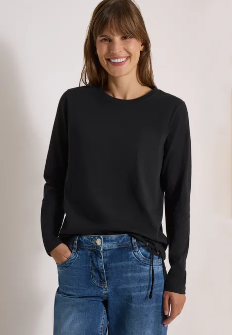 Lächelnde Frau trägt schwarzen Langarmpulli und blaue Jeans, Hände in den Taschen.