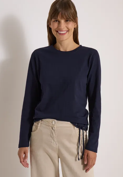 Eine lächelnde Frau in dunkelblauem Langarmshirt und beigefarbenen Hosen vor hellem Hintergrund.