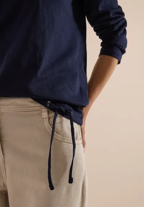 Eine Person in Marineblauem Langarmshirt und cremefarbenen Hosen mit Kordelzug, eine Hand in der Hosentasche.