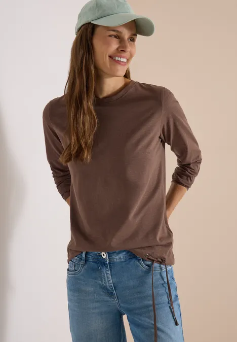 Junge Frau mit braunem Langarmshirt, Jeans und grüner Baseballkappe lächelt und posiert locker.