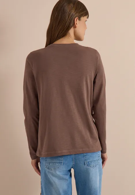 Rückansicht einer Frau in braunem Langarmshirt und blauen Jeans vor hellem Hintergrund.
