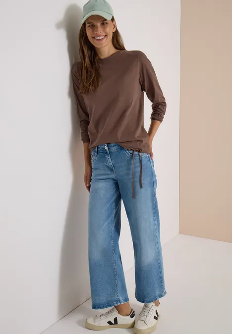 Eine Frau lehnt lächelnd an einer Wand, trägt braunes Langarmshirt, weite Jeans, weiße Sneaker und ein hellgrünes Cap.