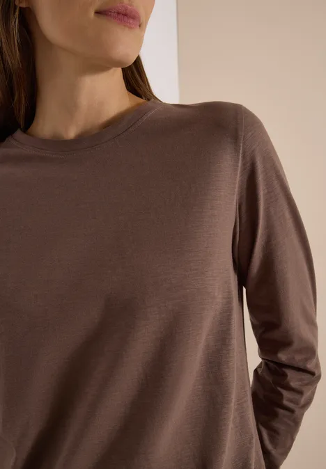 Nahaufnahme eines Oberkörpers in einem schlichten, braunen Langarmshirt vor neutralem Hintergrund.