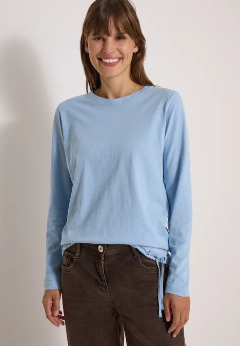 Eine lächelnde Frau in hellblauem Langarmshirt und braunen Cordhosen vor weißem Hintergrund.