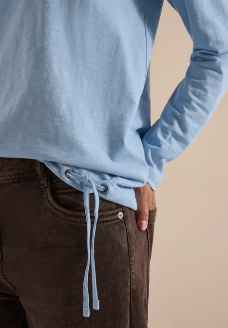Hellblauer Pullover mit Kordelzug am Bund, braune Hose, Hand in Tasche vor beigem Hintergrund.