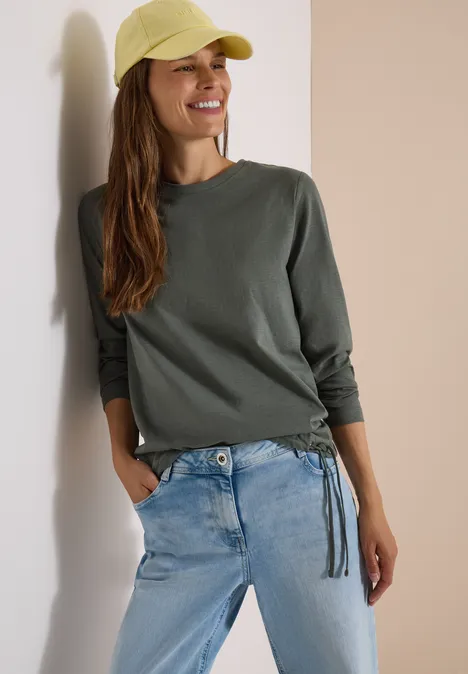 Eine lächelnde Frau trägt ein grünes Langarmshirt, helle Jeans und eine gelbe Baseballkappe, lehnt an der Wand.