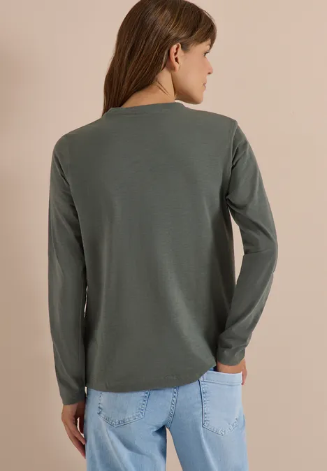 Rückansicht einer Frau in olivgrünem Langarmshirt und hellblauen Jeans vor neutralem Hintergrund.
