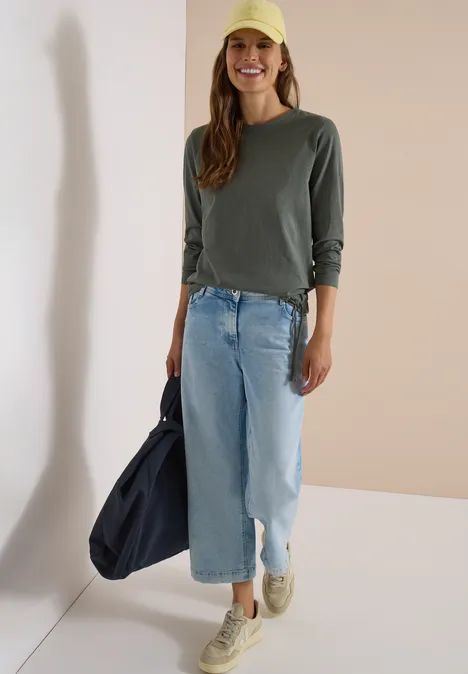 Frau mit grünem Langarmshirt, hellblauer Jeans, gelber Kappe, Sneakern und dunkelblauer Tasche vor Wand.