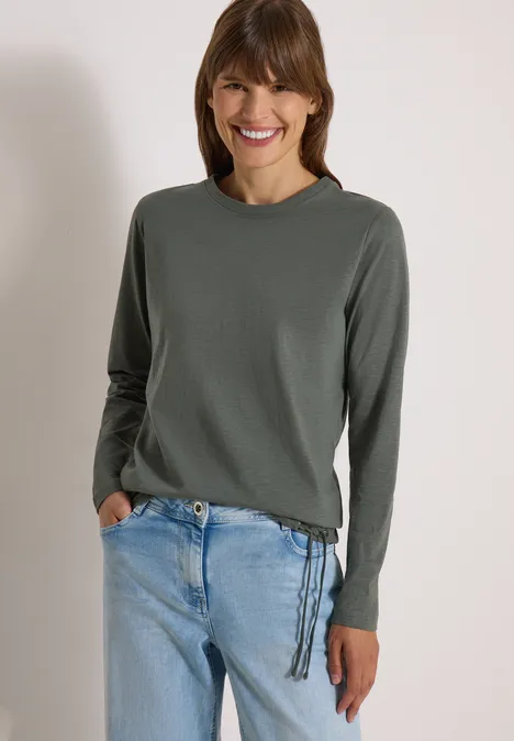 Frau lächelt in grauem, seitlich geschnürtem Pullover und hellblauen Jeans.