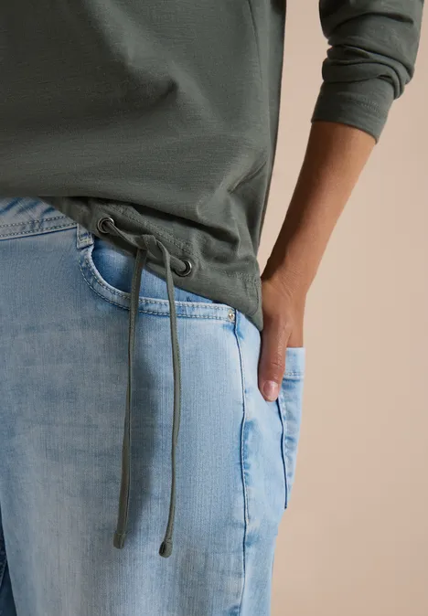Nahaufnahme einer Person in grüner Kapuzenjacke mit Kordelzug und hellblauen Jeans, eine Hand in der Tasche.