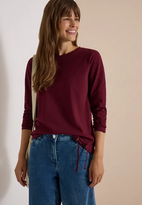Eine lächelnde Frau trägt eine kastanienrote Langarmbluse mit Tunnelzug und blaue Jeans vor hellem Hintergrund.