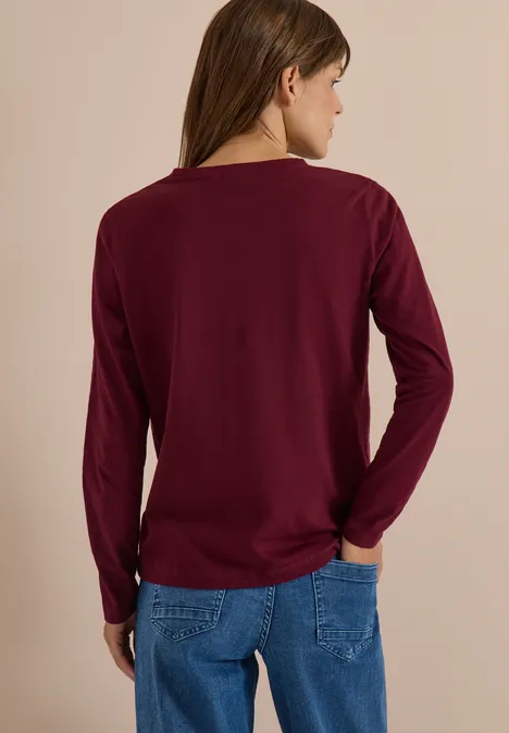 Eine von hinten fotografierte Person in einem dunkelroten Langarmshirt und blauen Jeans vor neutralem Hintergrund.
