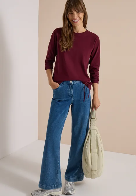 Frau in weitem Jeans-Hose und bordeauxrotem Pullover, lächelnd mit Tasche in der Hand.