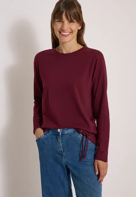Lächelnde Frau in burgunderrotem Langarmshirt und blauen Jeans vor neutralem Hintergrund.
