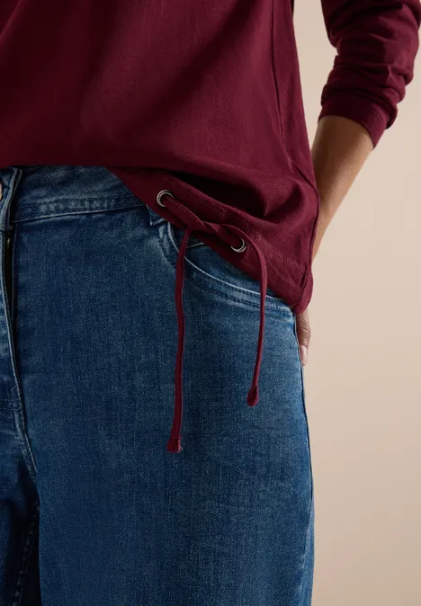 Person trägt dunkelrotes Shirt und weite Jeans mit Kordelzug am Bund.