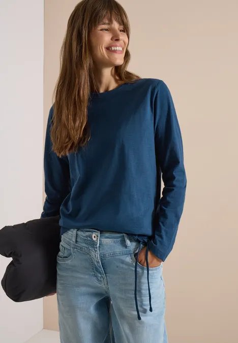 Langarm Shirt mit Rundhals und Tunnelzug moonlight blue Eine Frau in blauem Langarmshirt und Jeans lehnt lächelnd an einer Wand, hält ein Kissen in der Hand.