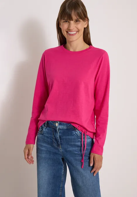 Langarm Shirt mit Rundhals und Tunnelzug pink flash Frau steht vor hellem Hintergrund, trägt pinken Pullover und blaue Jeans und lächelt in die Kamera.