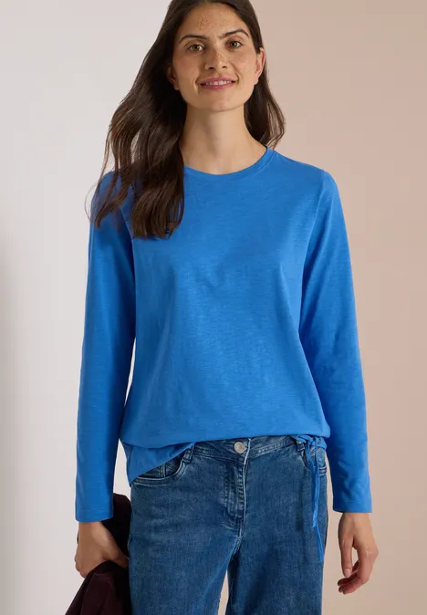 Een vrouw glimlacht voor een lichte achtergrond, ze draagt een blauw trui en spijkerbroek.