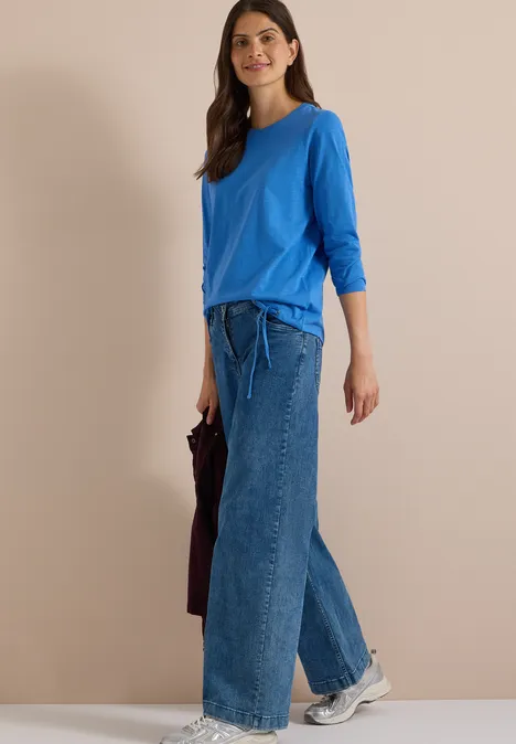 Vrouw in blauwe longsleeve en wijdvallende denimrok, witte sneakers, houdt een donkere tas vast.