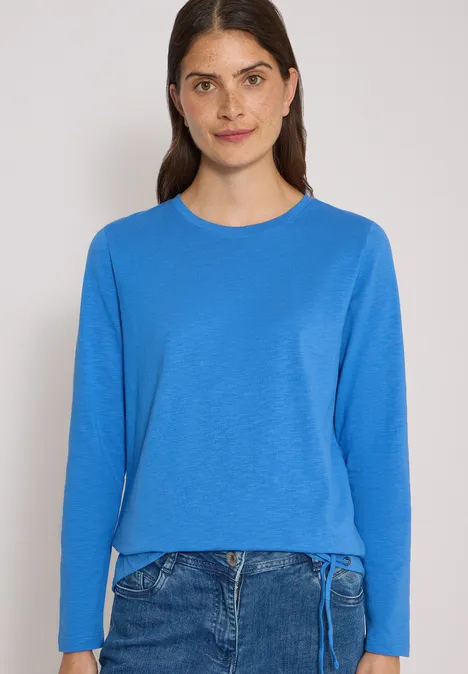 Vrouw in blauw shirt met lange mouwen en spijkerbroek voor effen achtergrond.