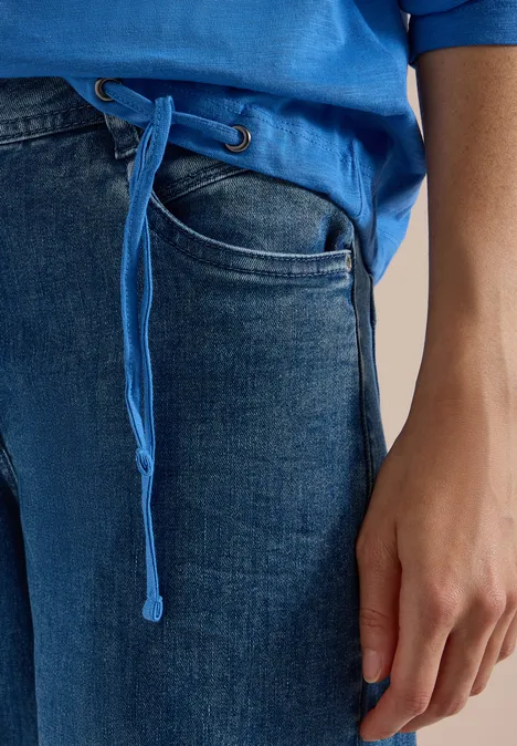 Close-up van een persoon in blauwe jeans en een blauw shirt met koord, hand langs het lichaam.
