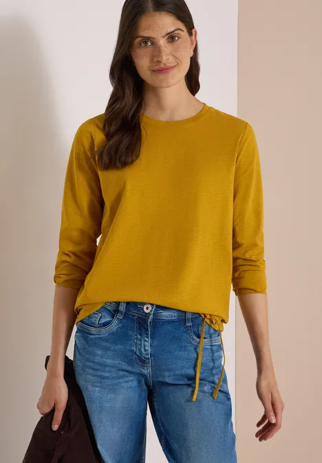 Femme en pull jaune à manches longues et jean bleu, tenant un sac noir.