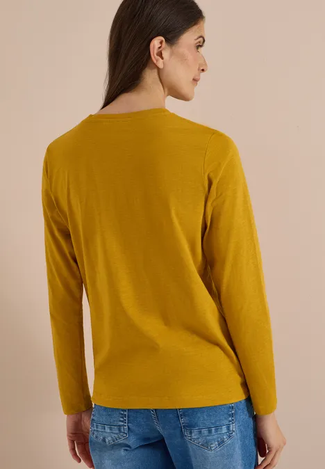 Vue de dos d'une femme portant un t-shirt à manches longues moutarde sur fond neutre.