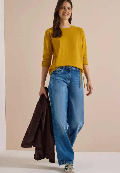 Femme en pull moutarde et jean large, tenant un manteau d'une main.