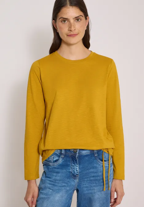 Femme aux cheveux foncés portant un pull moutarde et un jean devant un fond neutre.