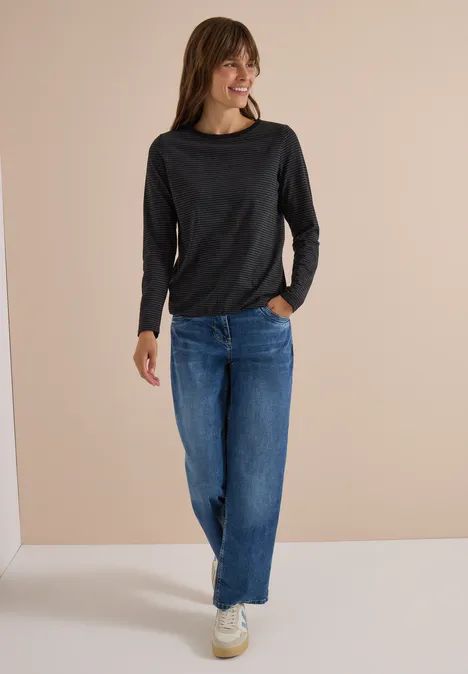 Frau trägt schwarzes Langarmshirt und weite blaue Jeans, lächelnd vor neutralem Hintergrund.