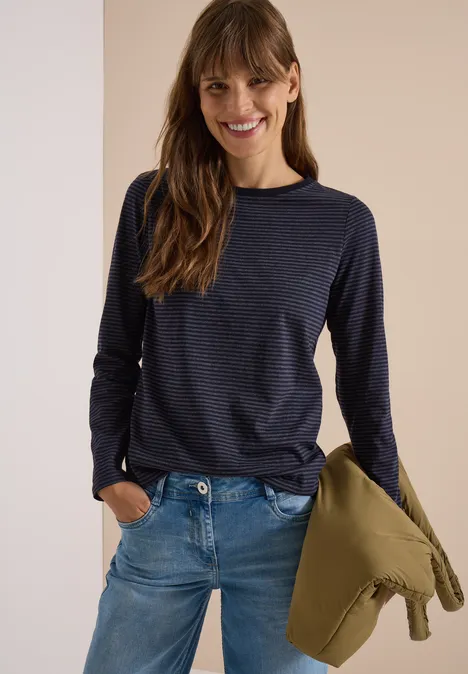 Frau in Jeans und gestreiftem Pullover hält Jacke, lächelt.