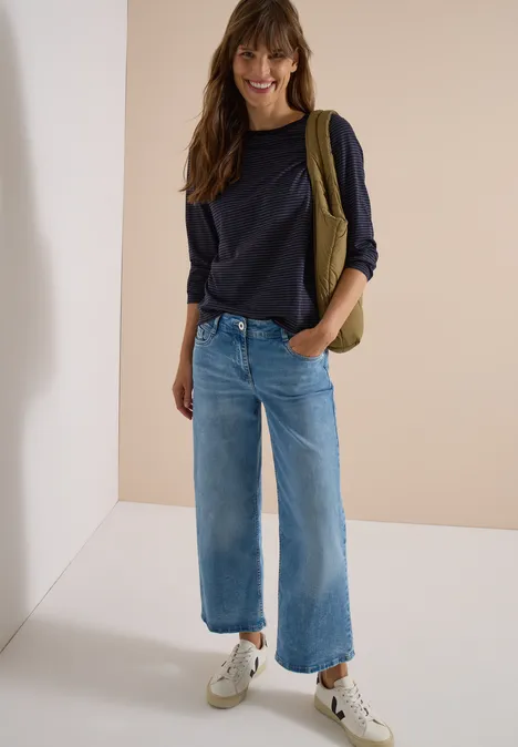 Fröhliche Frau mit dunkelblauem Pullover, weiten Jeans und Sneakers posiert vor schlichtem Hintergrund.