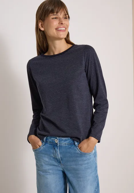 Frau in marineblauem Longsleeve und Jeans steht vor hellem Hintergrund und lächelt.