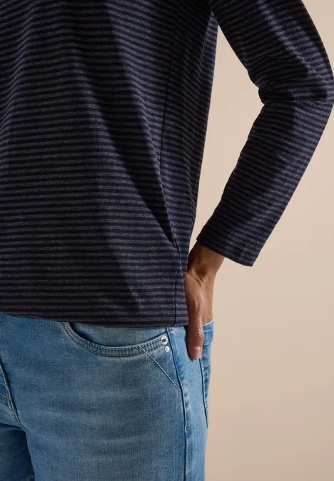 Teilansicht einer Person in gestreiftem Langarmshirt und Jeans, die Hand in der Tasche.