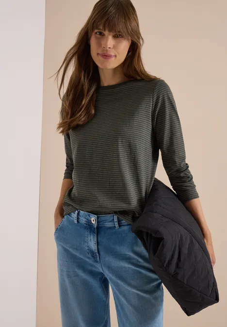 Junge Frau trägt grünes Langarmshirt und blaue Jeans, hält schwarze Jacke in der Hand.
