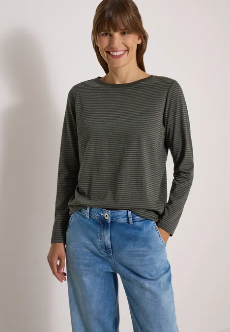 Eine lächelnde Frau trägt ein olivgrünes Langarmshirt und locker sitzende blaue Jeans, eine Hand in der Tasche.