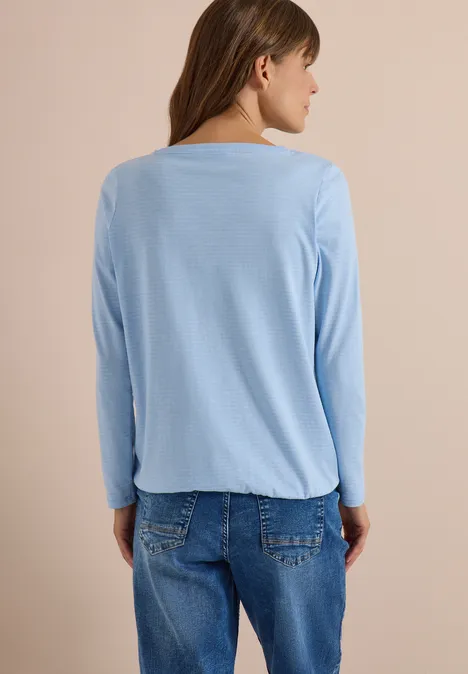 Frau von hinten in hellblauem Langarmshirt und Jeans vor neutralem Hintergrund.