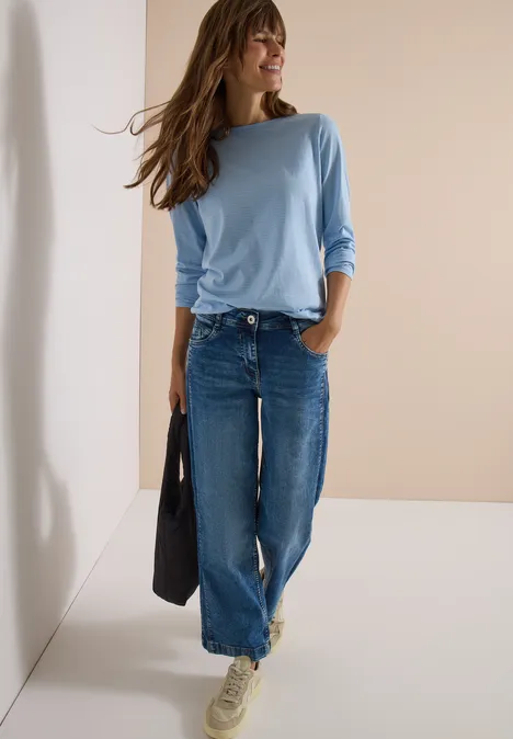 Frau in hellem Langarmshirt und weiten Jeans mit schwarzer Tasche vor neutralem Hintergrund.