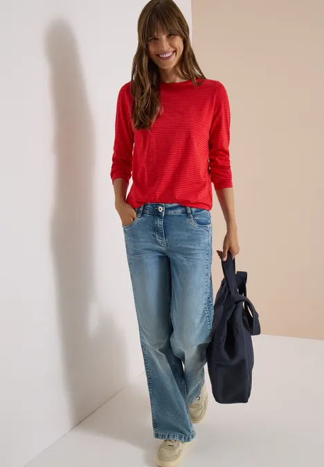 Lächelnde Frau in rotem Pullover und weiten Jeans, lehnt an weißer Wand und hält eine dunkelblaue Tasche.