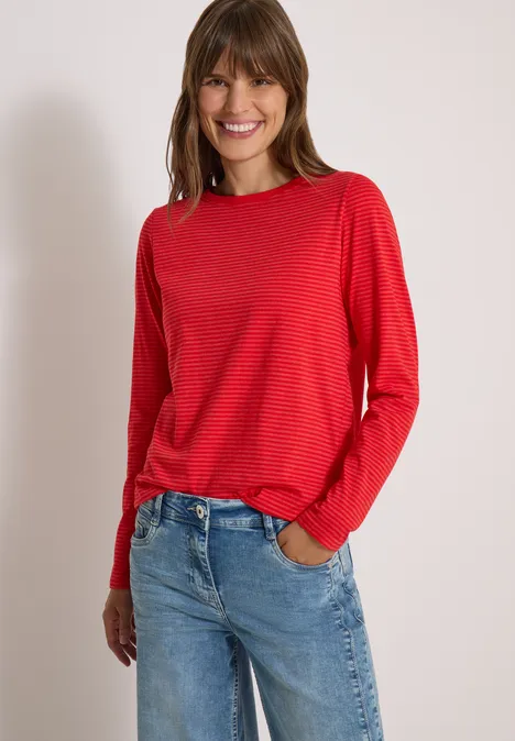 Frau lächelt, trägt rotes Langarmshirt und blaue Jeans vor weißem Hintergrund.
