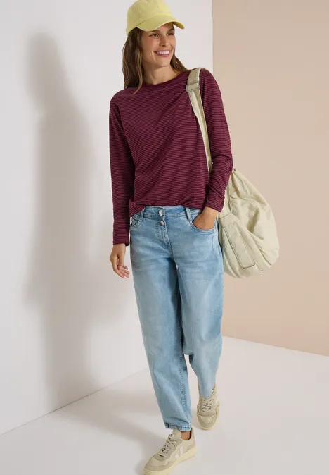 Frau mit gelber Kappe, weinrotem Pullover, hellblauen Jeans und großer beiger Tasche lehnt an hellem Hintergrund.