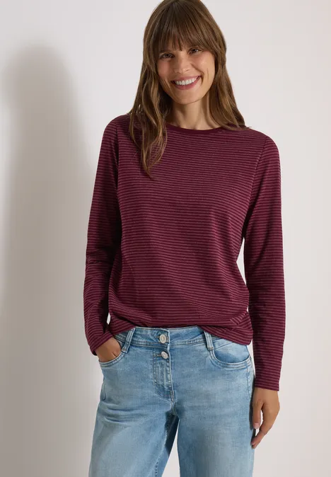 Frau in lockerem dunkelrotem Langarmshirt und Jeans, steht lächelnd mit Händen in den Taschen.