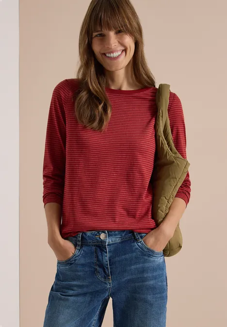 Langarm Shirt mit Boat Neck und Streifen russet red Lächelnde Frau in rotem Langarmshirt, blauen Jeans und olivgrüner Umhängetasche vor neutralem Hintergrund.